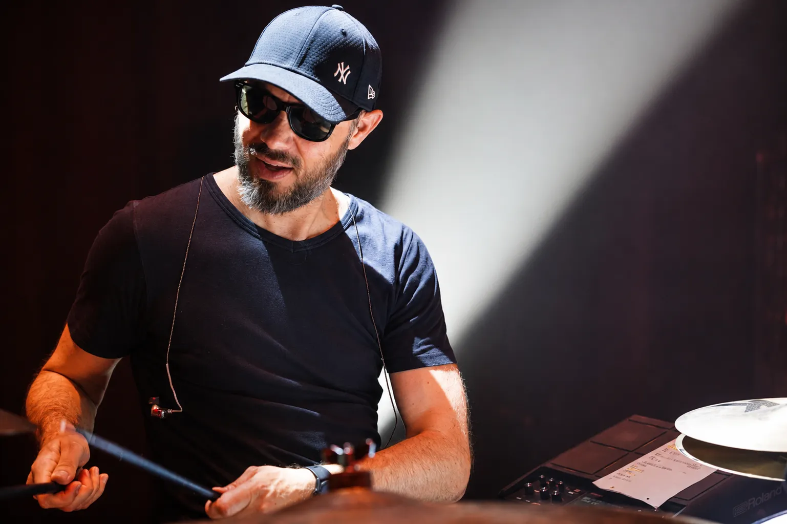 Le batteur de Victoria Orka, casquette et lunettes noires, baguettes en main derrière ses cymbales.