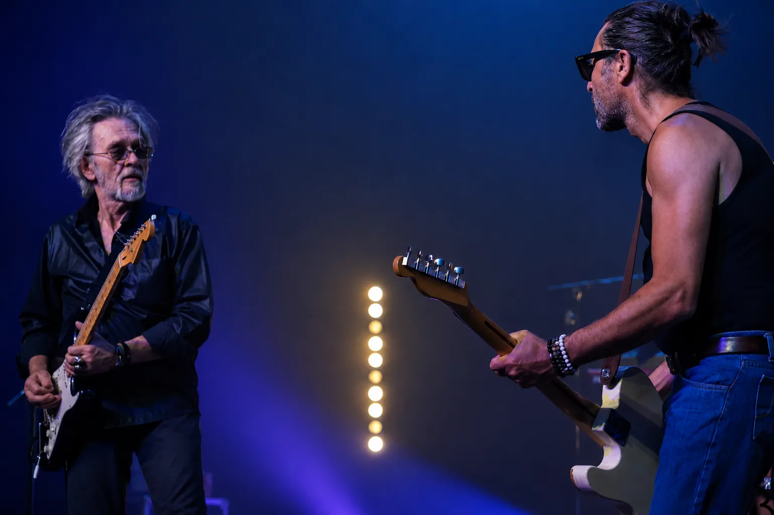 Deux guitaristes de Victoria Orka face à face sur scène, encadrant une rangée de spots dorés.