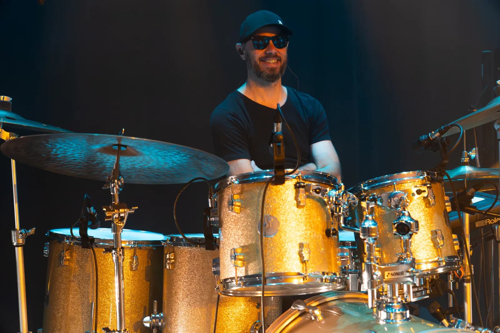 Le batteur de Victoria Orka souriant derrière un kit scintillant doré, lumière chaude de scène.