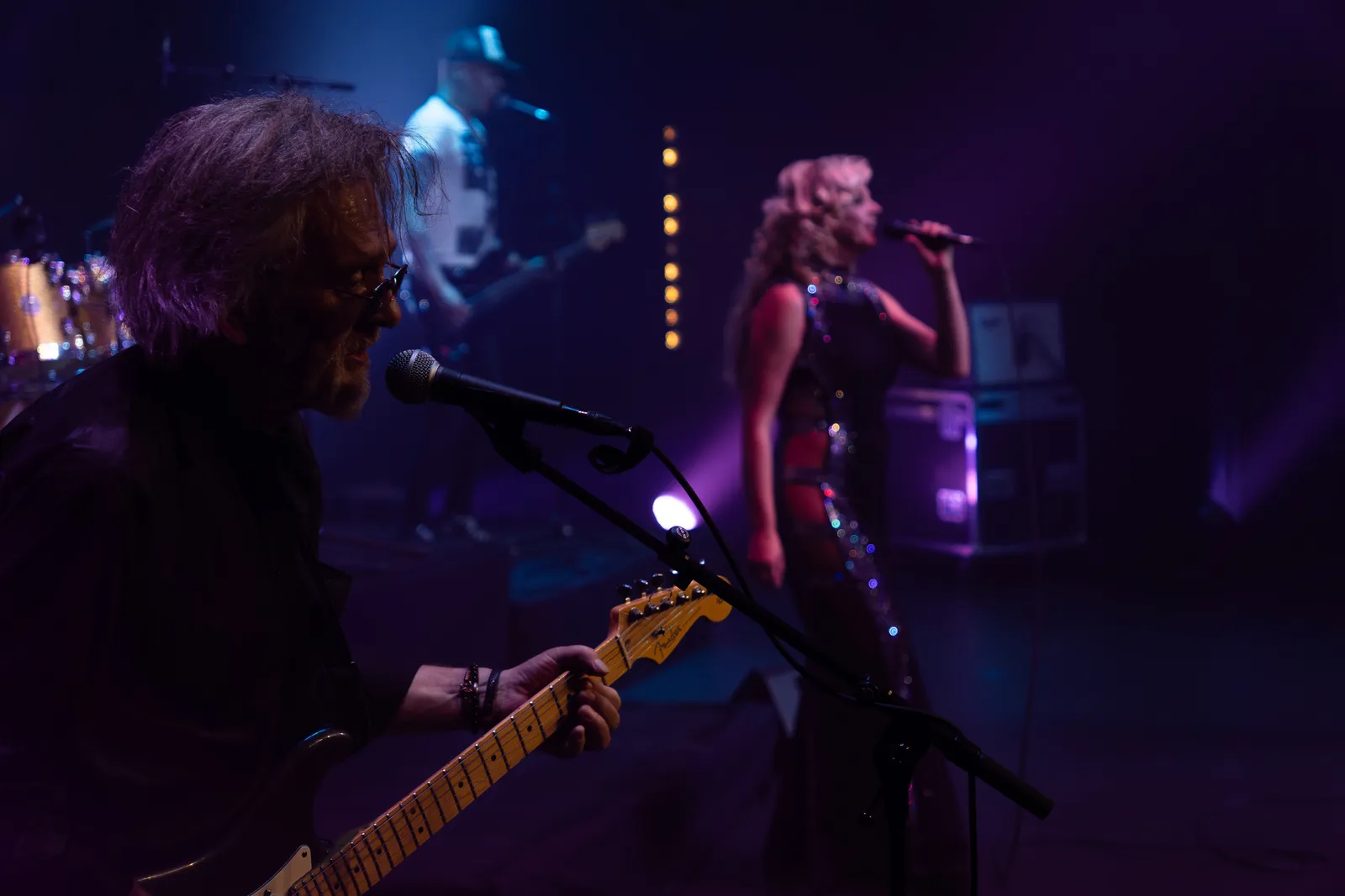 La chanteuse de Victoria Orka au micro en robe pailletée, guitariste et bassiste en amorce au premier plan.
