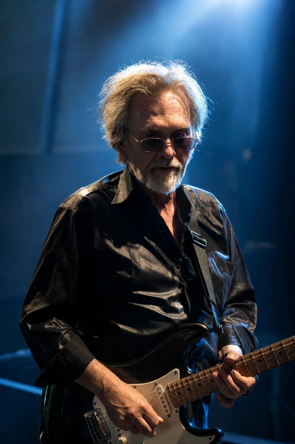 Portrait du guitariste de Victoria Orka, cheveux blancs et lunettes teintées, sous une lumière bleu nuit.